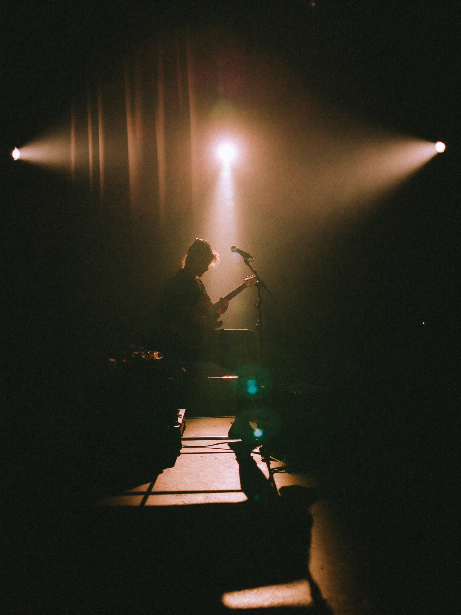 An artist backstage tuning a guitar before a show, stage light leaking through the curtain