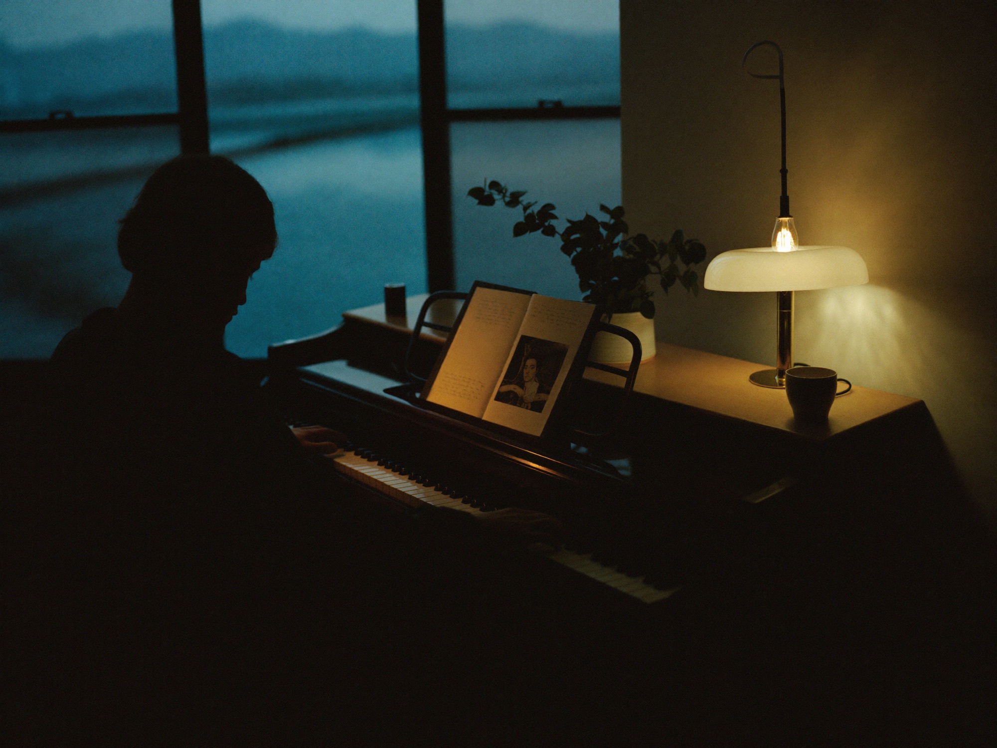 A songwriter at an upright piano in warm lamplight at dusk