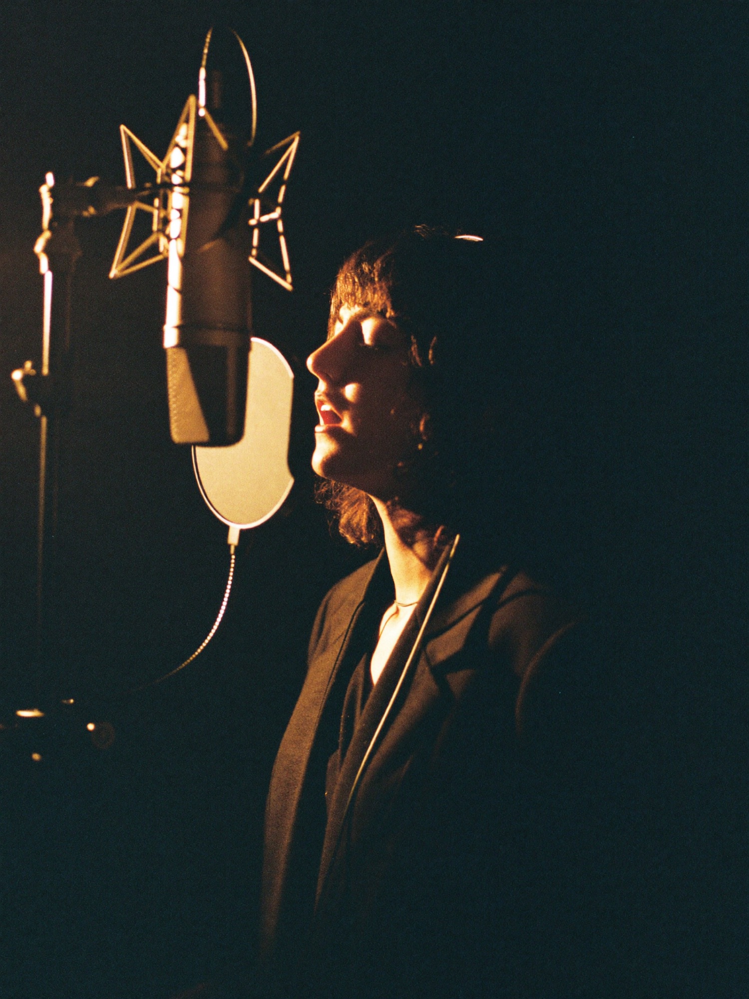 A vocalist singing into a condenser microphone in a dim booth with warm rim light
